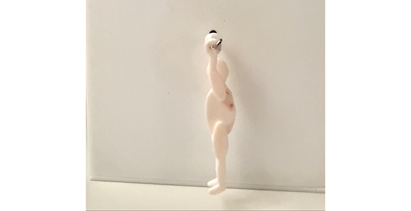 Seitenansicht einer kleinen, hellen menschlichen Figur vor weißem Hintergrund. Im Gegensatz zum vorherigen Bild hängt die Figur aufrecht an einer Befestigung am Kopf, während ihre Füße frei über dem Boden schweben.